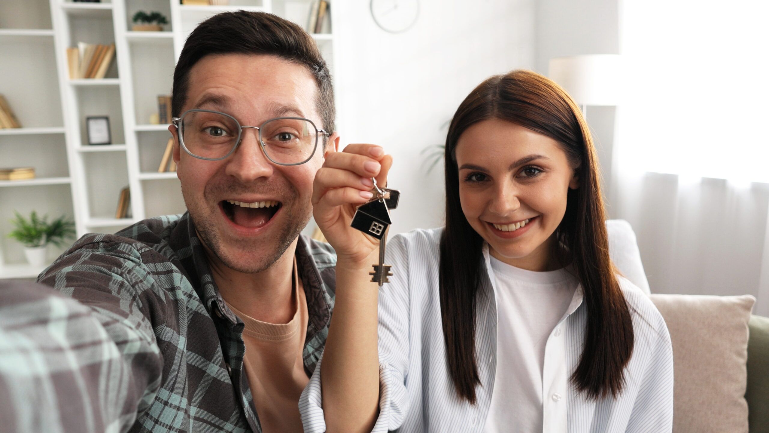 The first house. Happy couple, new home buyers, looking at phone camera while making video calls to relatives. Young couple celebrates moving into modern cottage, showing the house keys to the camera.